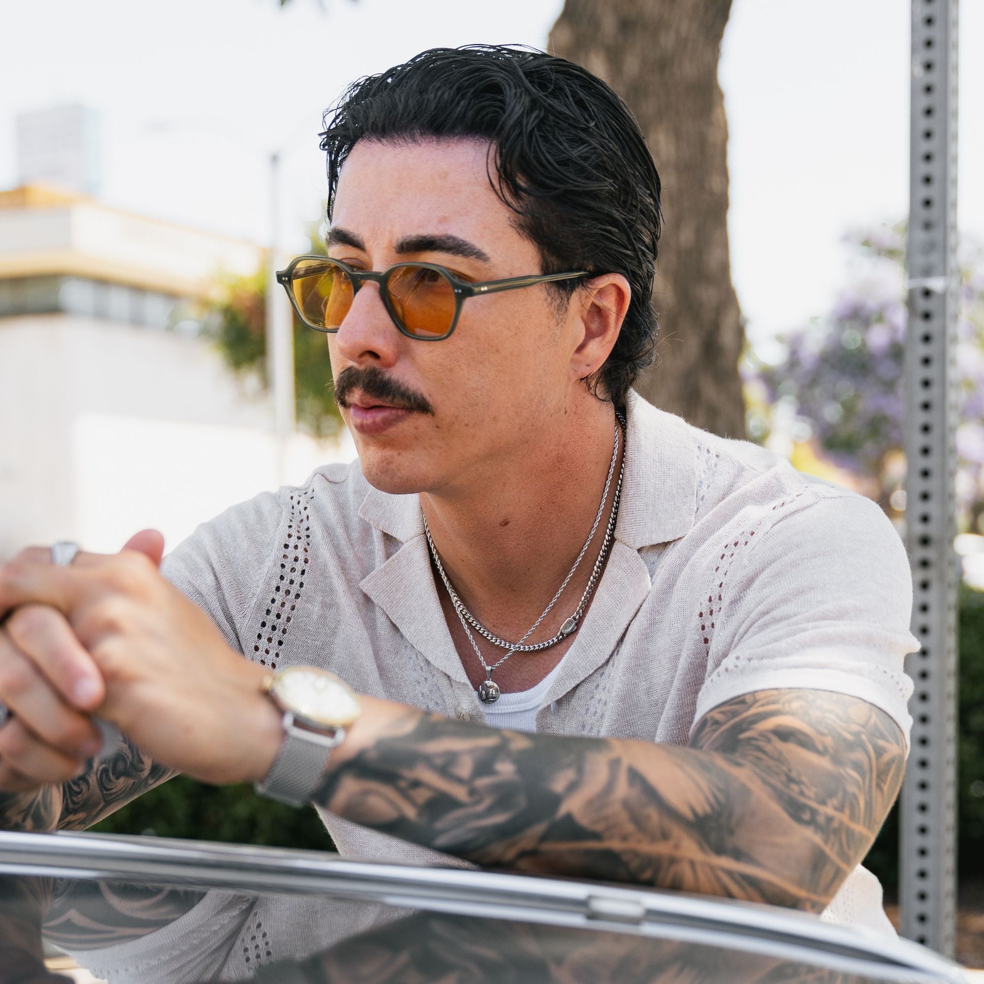 A man with a mustache and tattoos wearing orange-tinted sunglasses, a light-colored short-sleeve shirt, and layered silver necklaces, leaning on a car outdoors.
