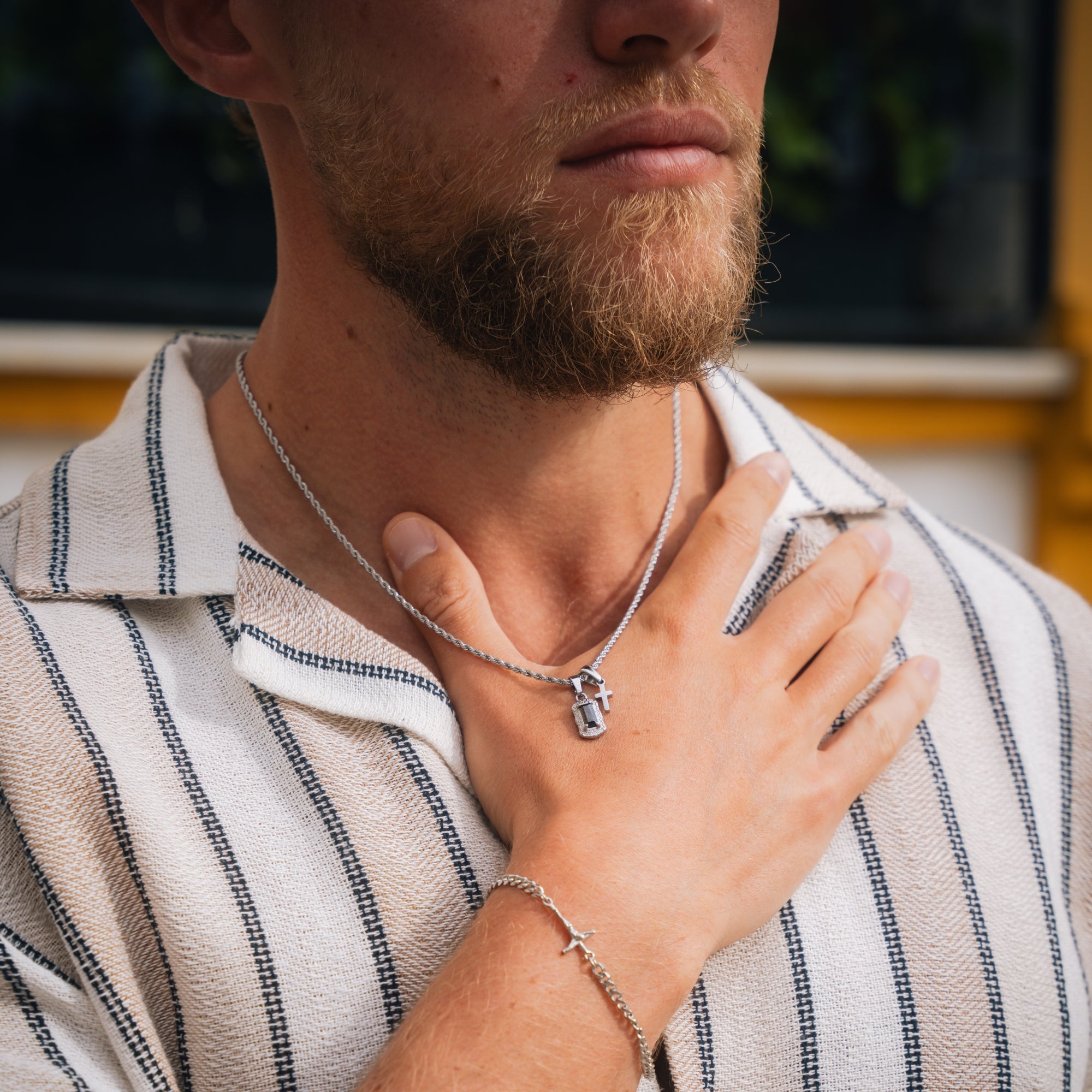 Man wearing a striped shirt with a necklace and bracelet, outdoors.