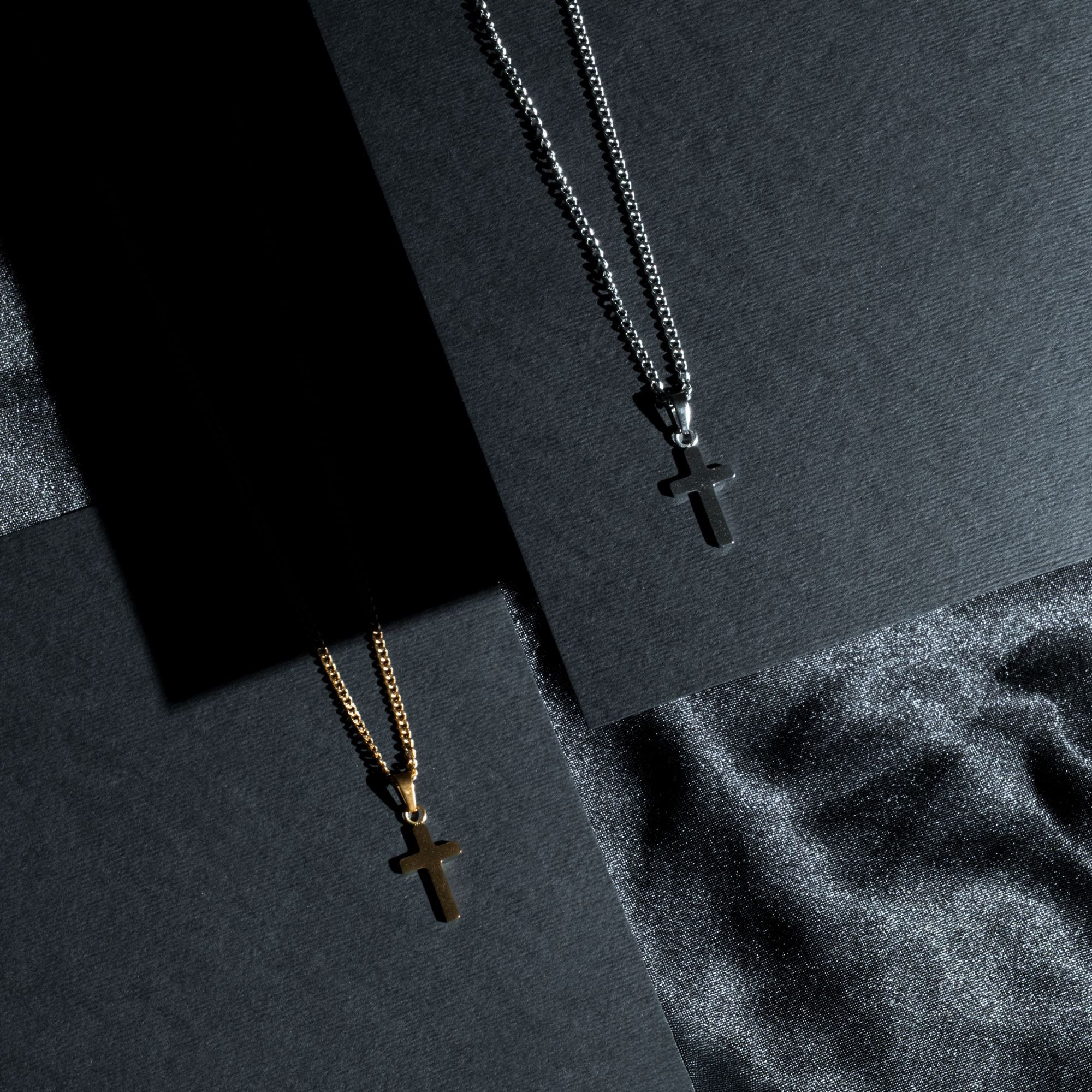 Two cross pendant necklaces displayed on dark textured cards and fabric. One necklace is gold-toned with a cross pendant on a gold chain, and the other is silver-toned with a cross pendant on a silver chain.
