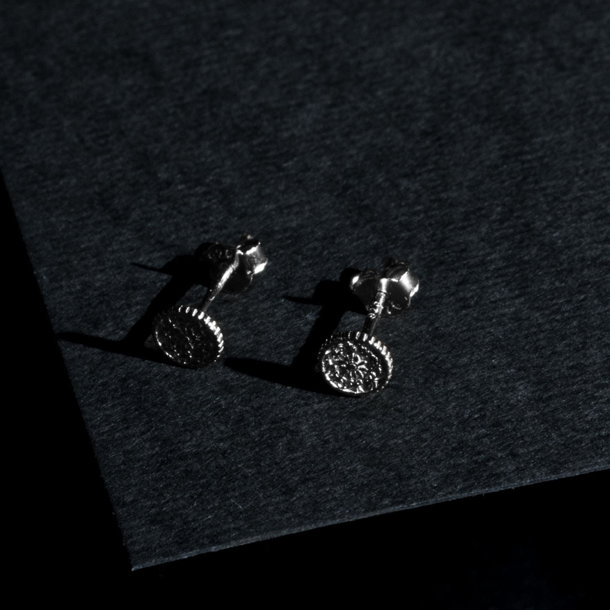 A pair of silver-toned coin stud earrings with textured circular faces, displayed on a dark textured surface.