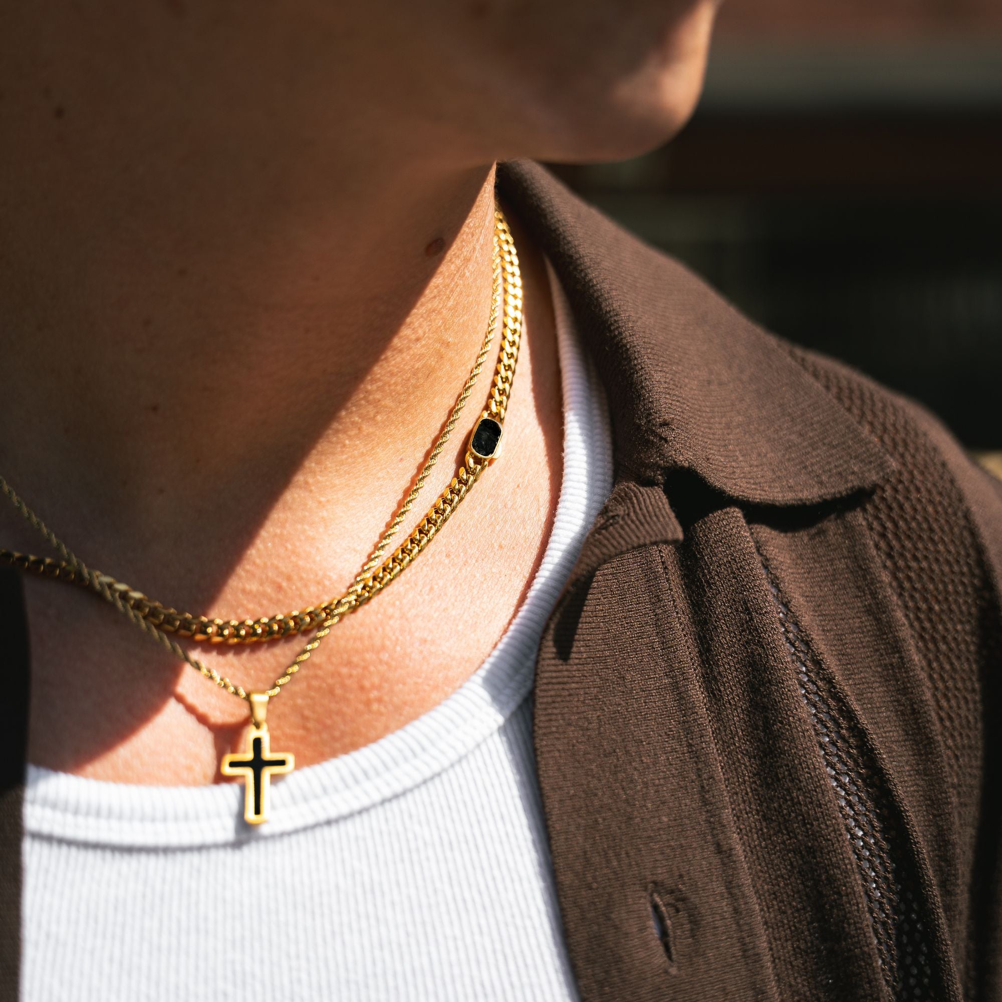 Close-up of a person wearing layered gold necklaces, including a cross pendant and a chain with a small black oval stone, over a white shirt and brown jacket.