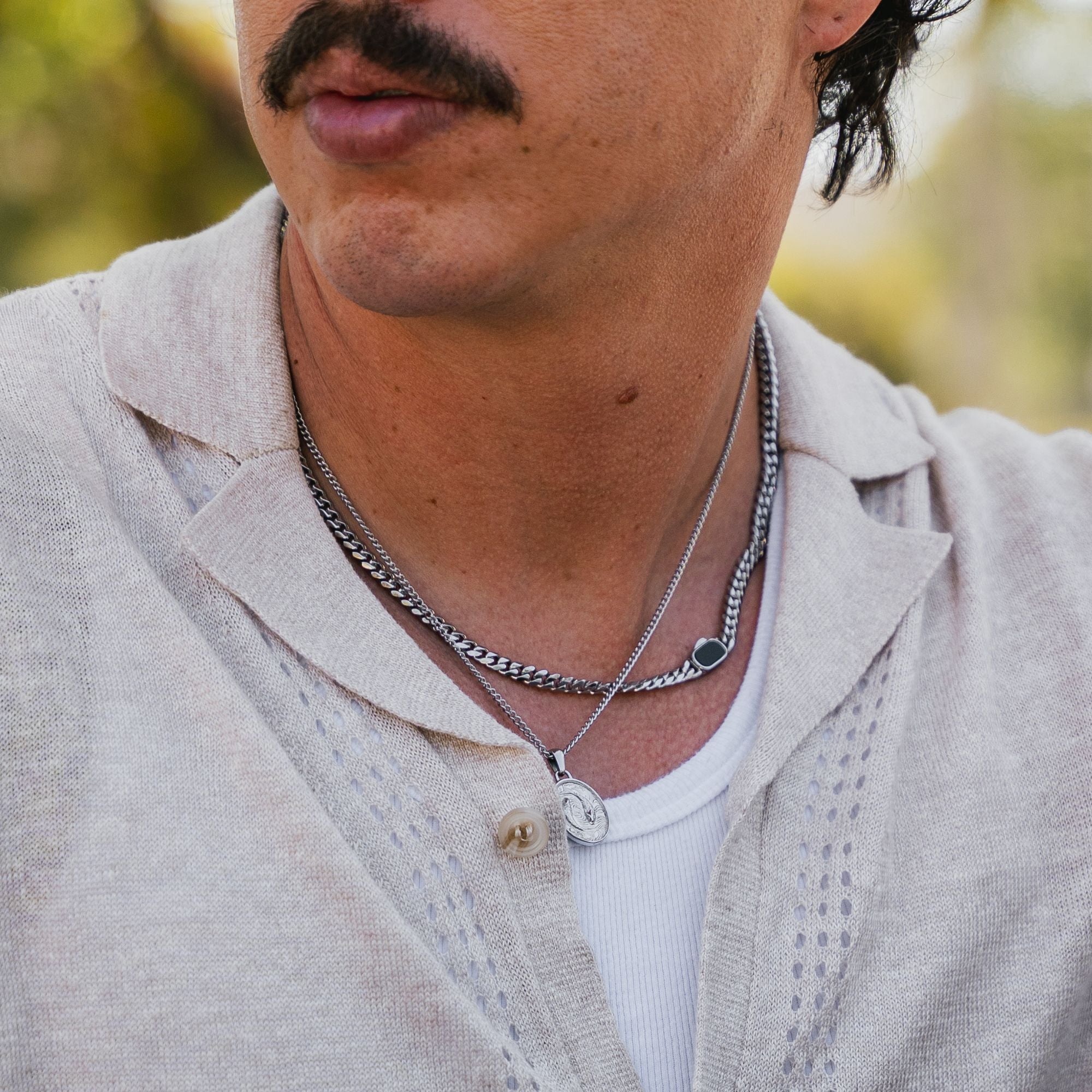 A close-up of a man wearing layered silver necklaces, including a curb chain necklace and a delicate chain with a round pendant, over a light beige knit polo shirt.