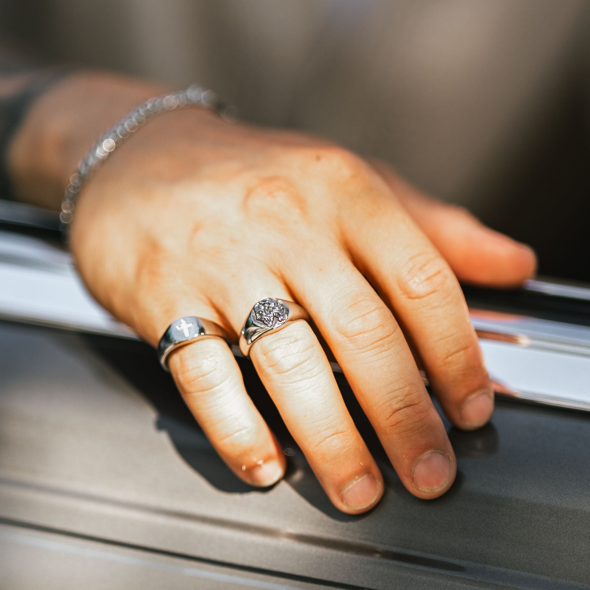 A close-up of a person's hand resting on a dark metallic surface, wearing two silver rings and a silver chain bracelet.
