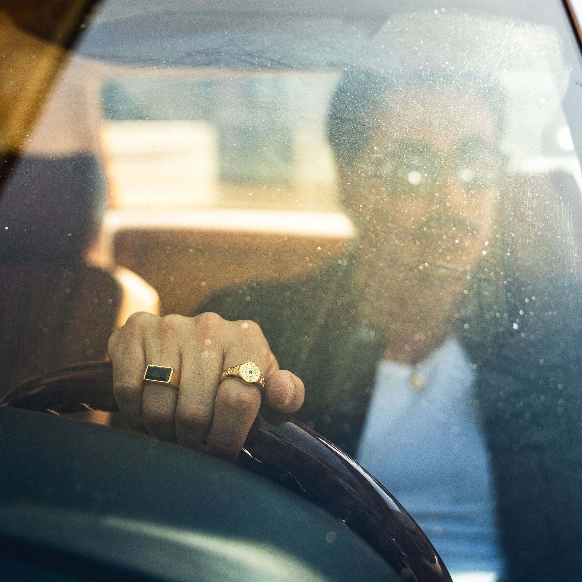 A close-up lifestyle shot of a man driving a car, wearing a gold signet ring with a black rectangular stone and a gold watch with a white dial on his wrist.