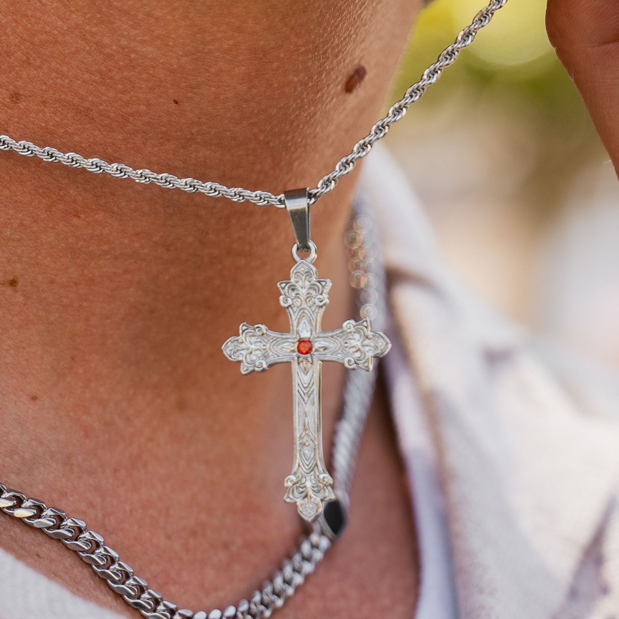 A close-up of a silver cross pendant necklace worn on a person's neck, featuring an ornate engraved design and a small red gemstone at the center, layered over a silver chain.
