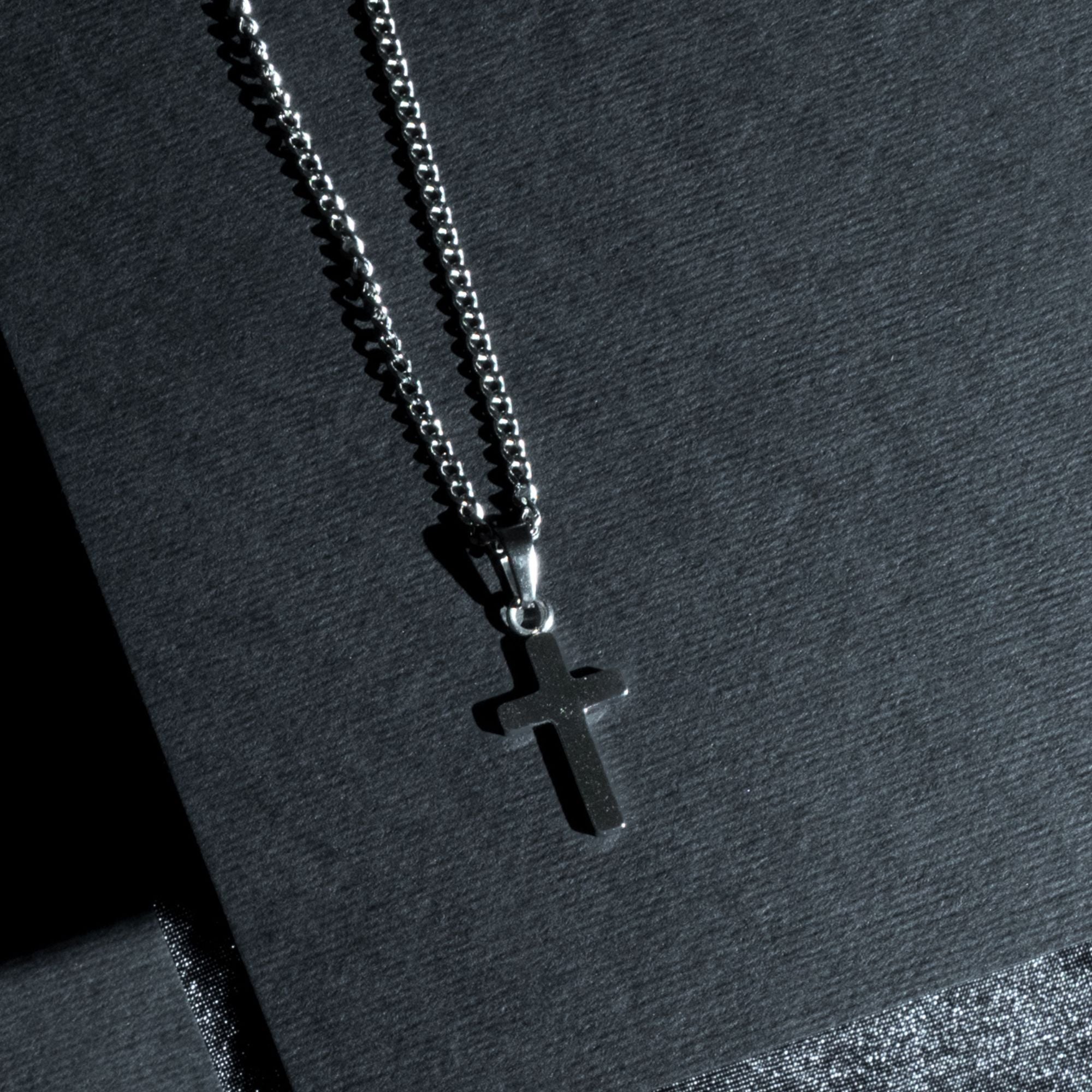 A silver-toned cross pendant necklace on a matching silver chain, displayed on a dark textured surface.