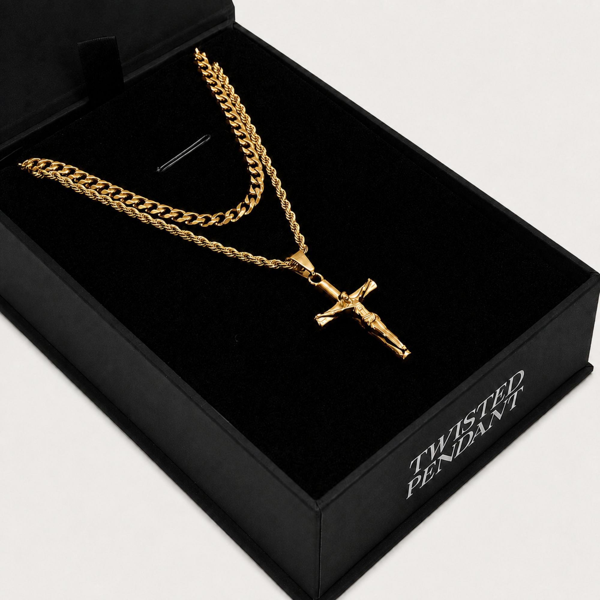 A gold-tone crucifix pendant necklace displayed inside an open black presentation box. The necklace features a thick curb chain and a smaller chain layer with a detailed crucifix pendant.
