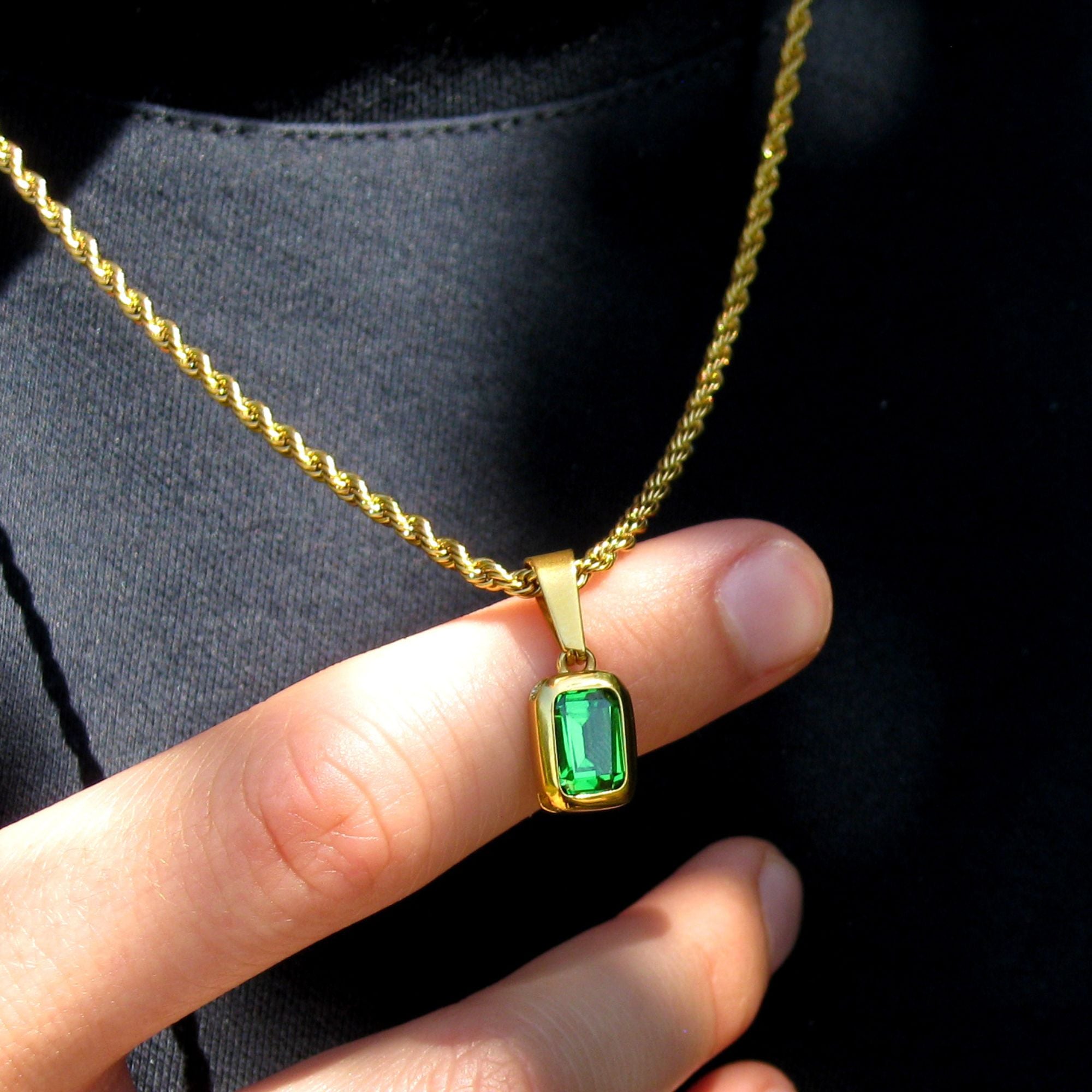 A gold rope chain necklace with a rectangular green gemstone pendant, held by a hand against a black shirt background.