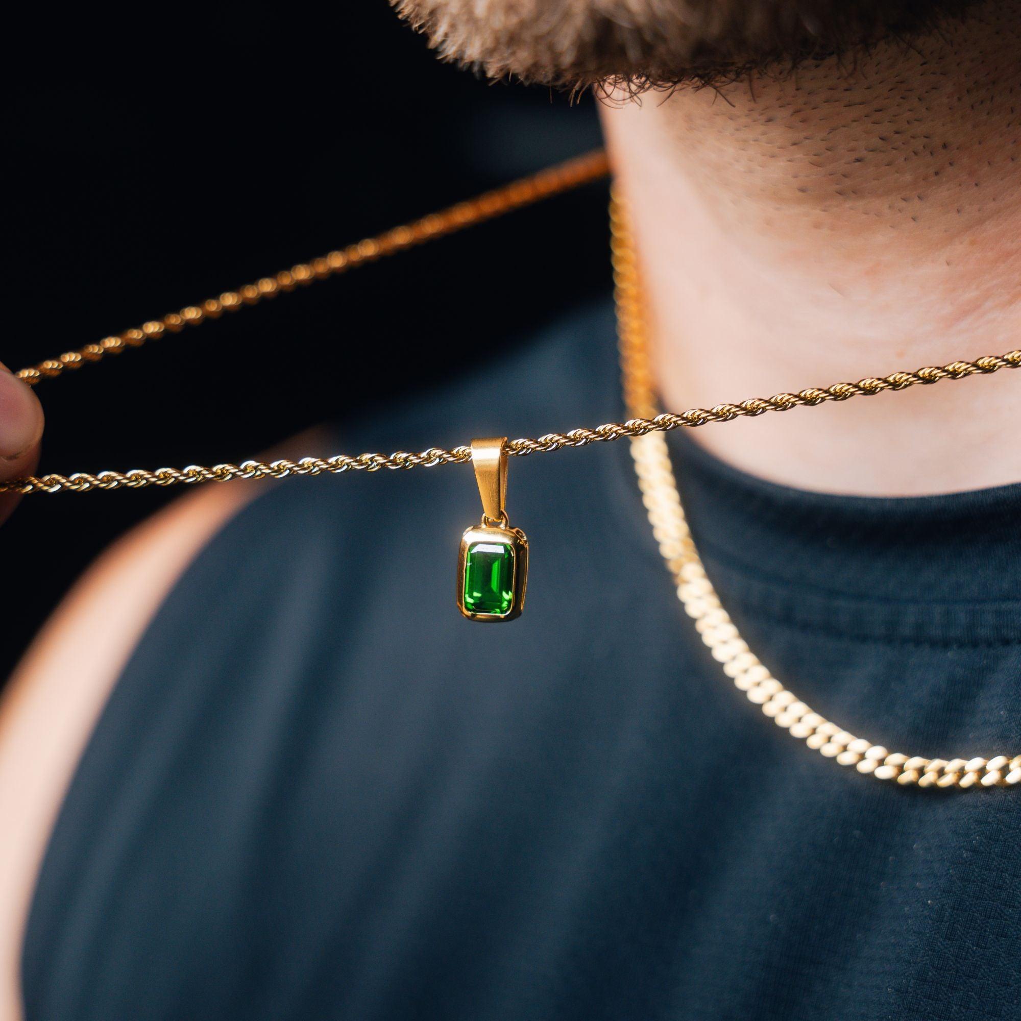 A close-up lifestyle shot of a man wearing a gold chain necklace with a rectangular green gemstone pendant, held by a hand against a dark background.