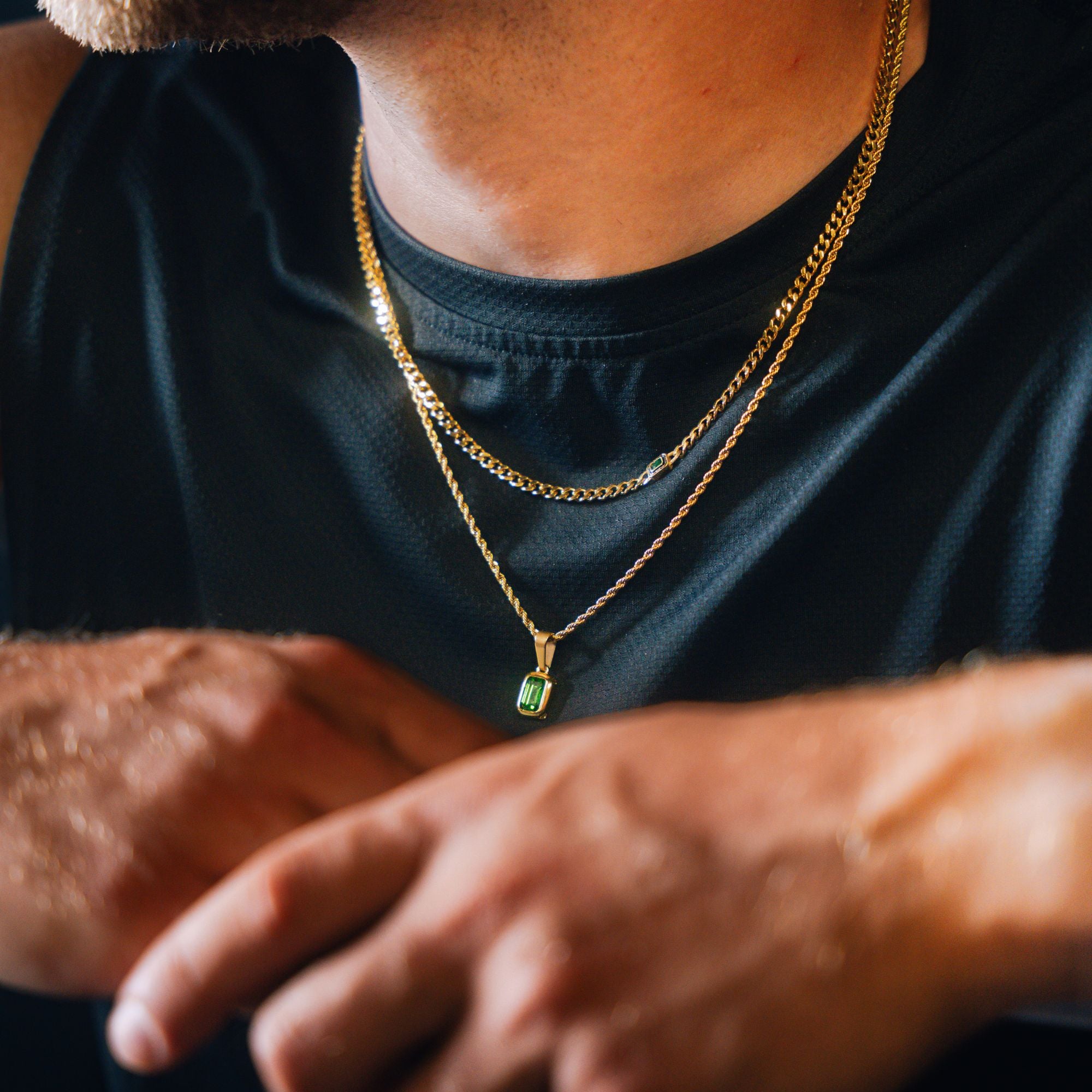 A close-up of a man wearing two gold necklaces, one with a small rectangular green pendant, over a black athletic shirt.