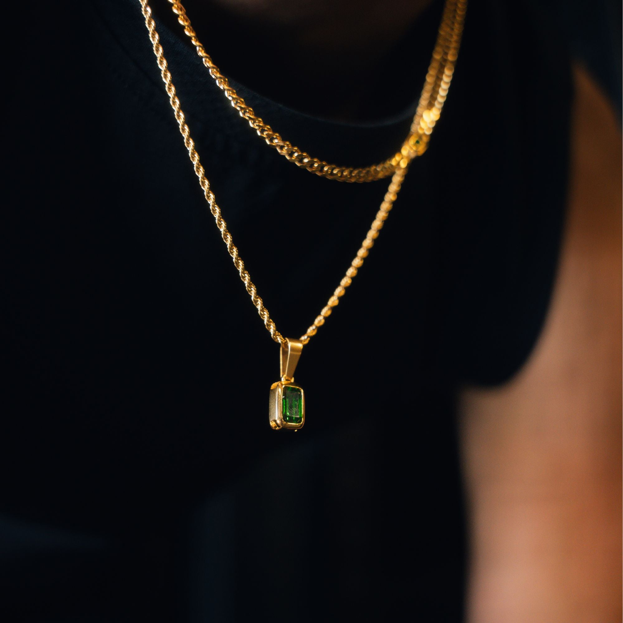 A close-up of a gold necklace with a rectangular green gemstone pendant, worn layered over a black shirt.
