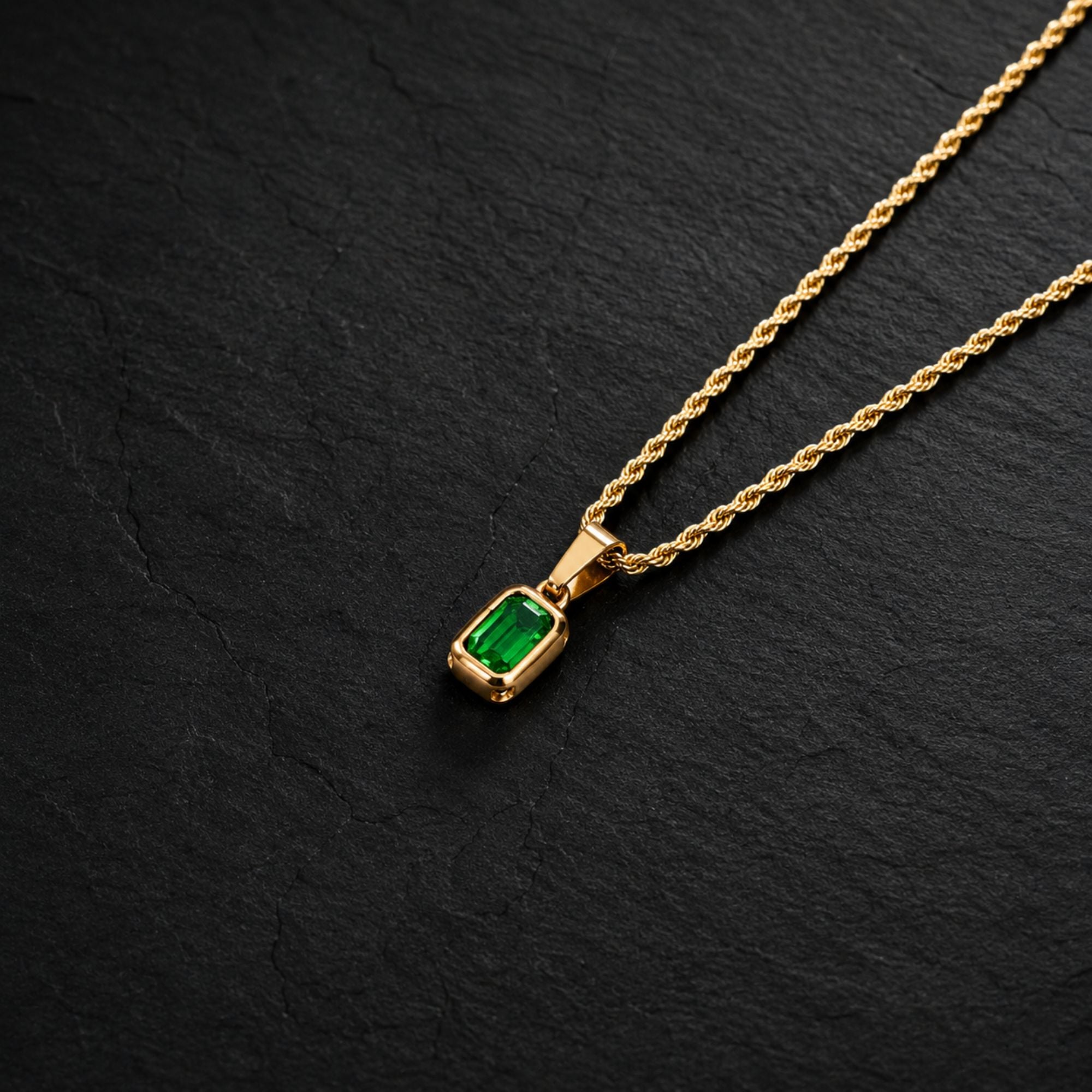 A gold chain necklace with a rectangular green emerald-cut gemstone pendant, displayed on a textured black slate background.