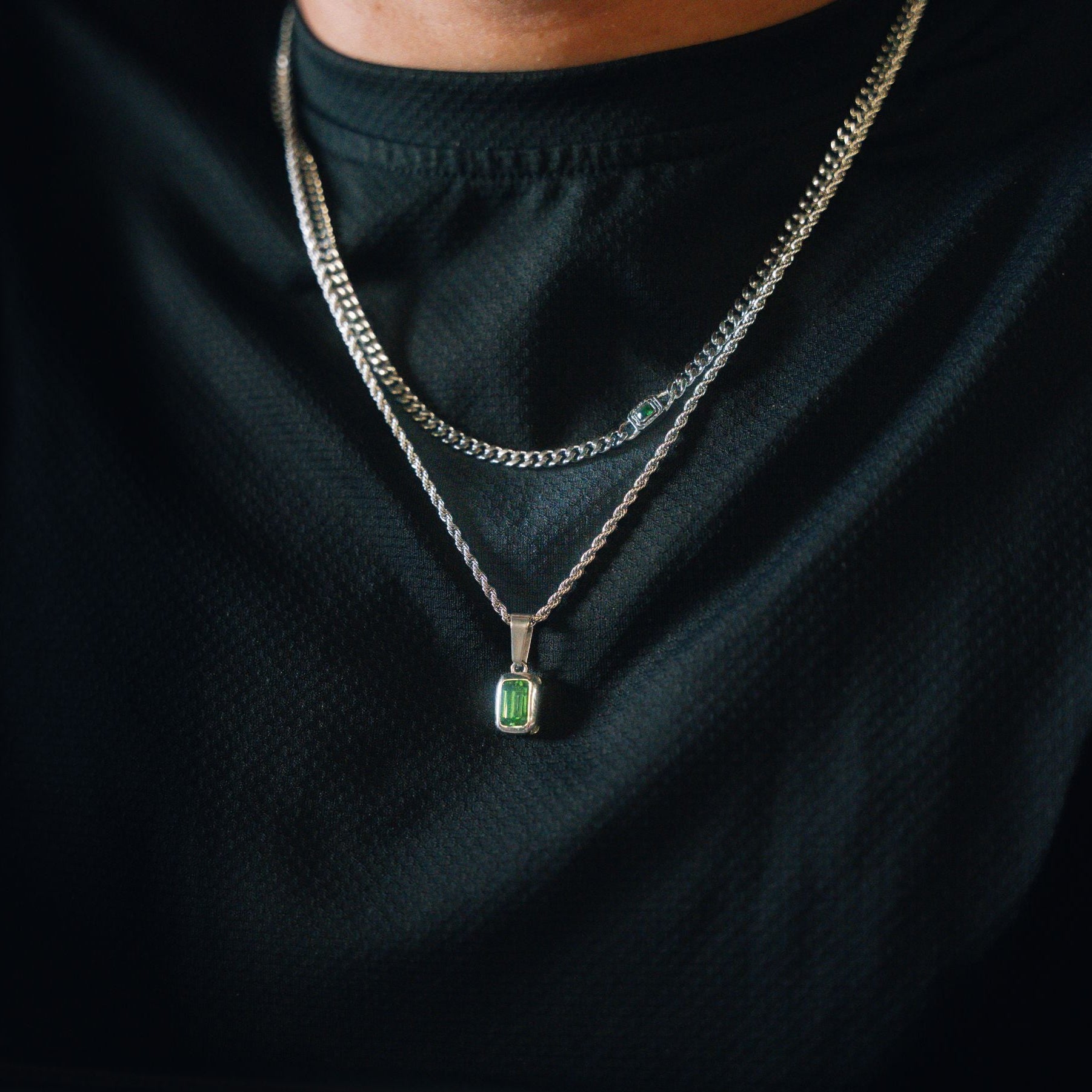 A close-up of a person wearing a silver-toned double-layer necklace with a rectangular green gemstone pendant, styled over a black textured t-shirt.