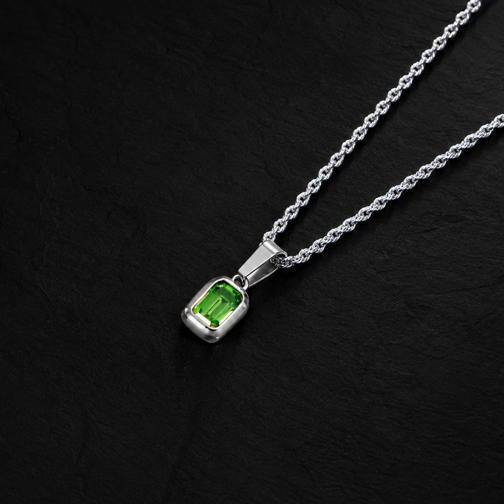 A silver necklace with a rectangular green gemstone pendant, displayed on a textured black background.