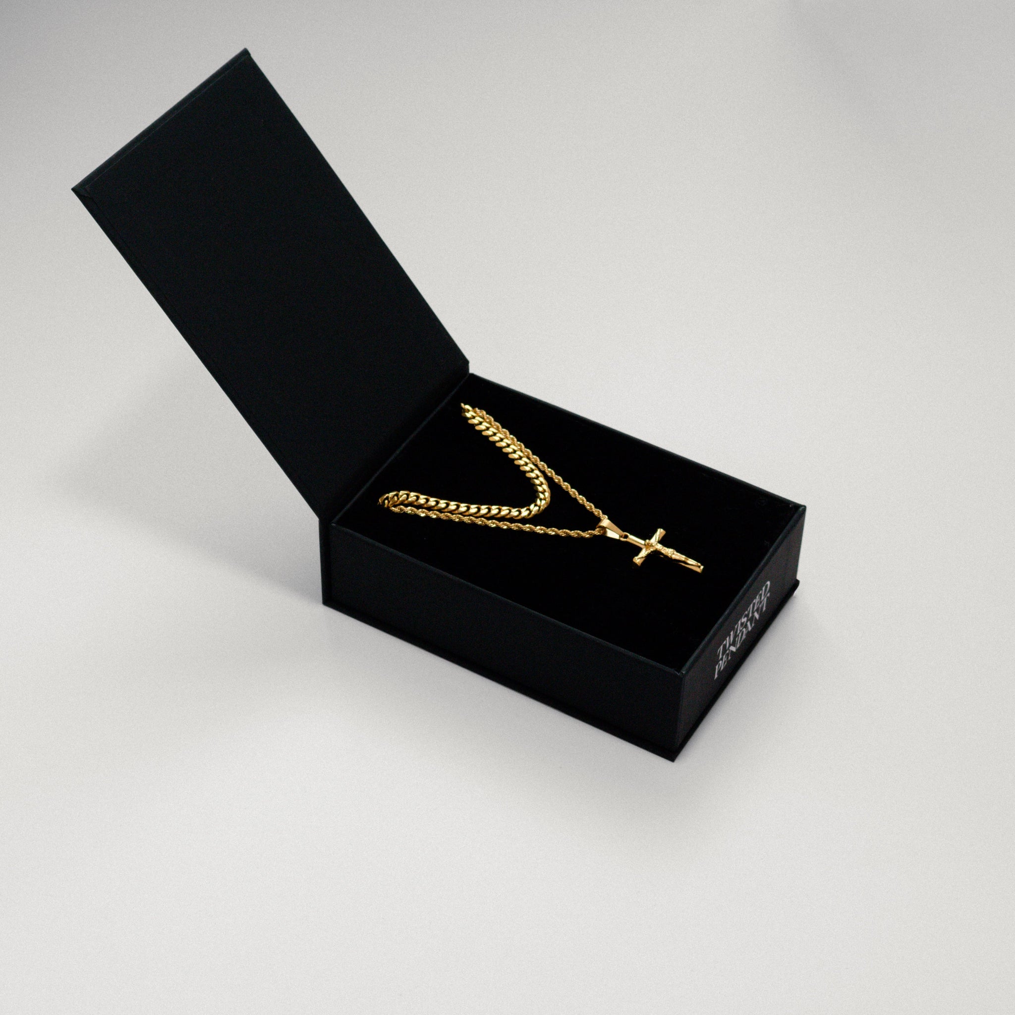 A gold cross pendant necklace displayed inside an open black presentation box on a light gray background.
